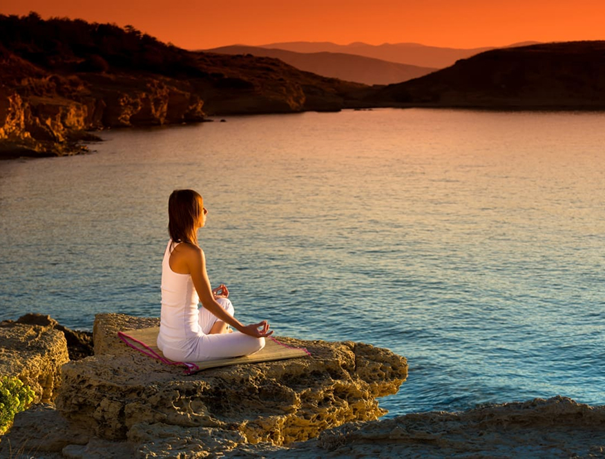 A woman meditating on a rock ledge above calm water at sunset