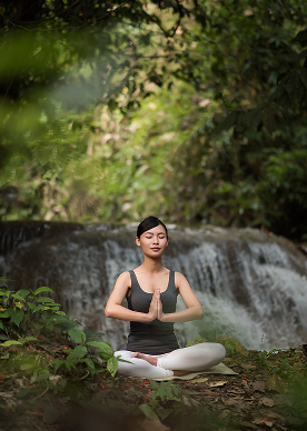 A woman is meditating in lotus pose with hands in prayer beside a forest waterfall
