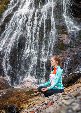 A woman seated in meditation with hands in gyan mudra in front of a rocky waterfall